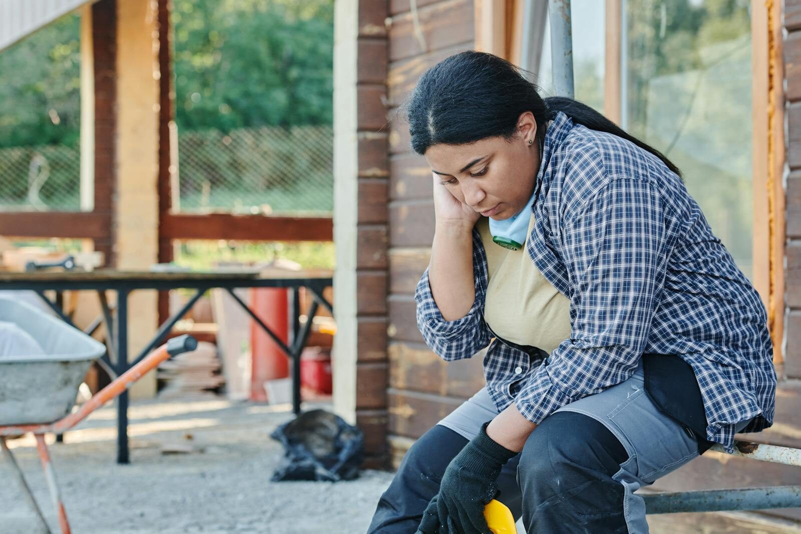 An African American woman in a plaid shirt sits, appearing tired at a construction site.